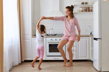 Dark haired little girl dancing with mother, daughter feeling amazing dancing with her loving mother in kitchen, family wearing casual style having fun together.