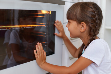 Closeup profile portrait of dark haired little cute charming female looking at oven, waiting when baking will be ready, child wearing white casual style t shirt.