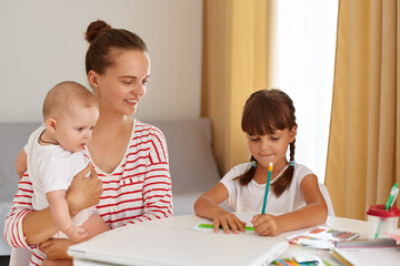 Smiling mother with new born baby in hands and elder daughter sitting at table and doing homework,...