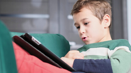 Cheerful little boy smiling while sitting on couch and using tablet at home. Modern kid and education technology. little boy looking at touch pad at home.