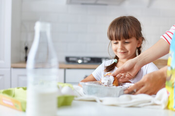 Obraz na płótnie Canvas Positive smiling female child with two pigtails standing near table, waiting her mom giving her hot tasty baking, kid girl wearing white t shirt posing in kitchen.
