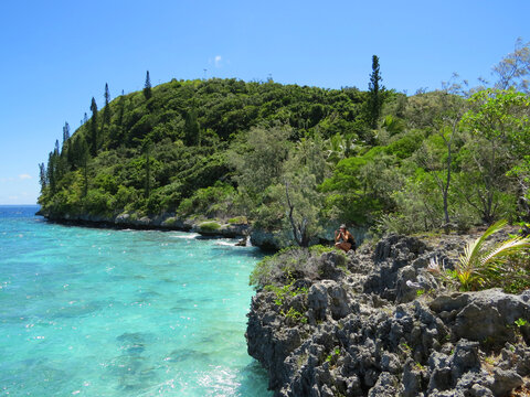 Caucasian woman taking a picture with the camera while squatting on a lush, exotic, green, tropical island with a rocky and rugged coastline terrain, next to a turquoise ocean.  