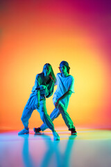 Young hip-hop dancers, stylish emotive girl and boy in action and motion in casual sports youth clothes on gradient multi colored background at dance hall in neon light.