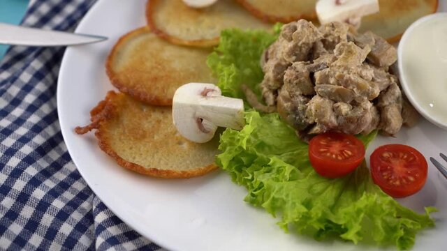 Deruny - potato pancakes with mushrooms, champignons sour cream on a white plate. Cherry tomatoes, salad. Restaurant serving on a blue background, checkered napkin, fork, knife. Pancake vegetable food