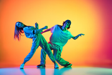 Two dancers, young girl and boy dancing hip-hop in casual sports youth clothes on gradient multi colored background at dance hall in neon light.