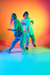 Young hip-hop dancers, stylish emotive girl and boy in action and motion in casual sports youth clothes on gradient multi colored background at dance hall in neon light.