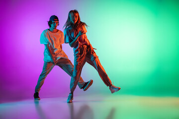 Stylish young hip-hop dancers, emotive girl and boy in action and motion in casual sports youth clothes on gradient multi colored background at dance hall in neon light.