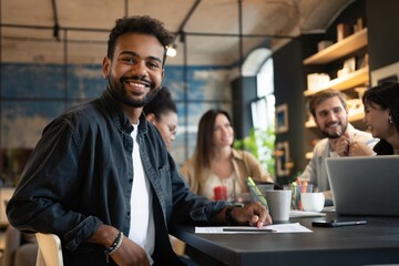 Portrait of confident mixed-race businessman sitting against colleagues in meeting room at office.