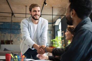 Two men shaking hands and looking at each other with smile while their coworkers sitting at the business meeting