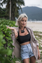 Fototapeta premium Beautiful caucasian young woman with blond hair, wearing casual clothing and stylish necklace. Standing at the beach. Looks directly at camera. 