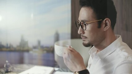 through glass view of Asian designer taking a coffee break in office walking and looking out window