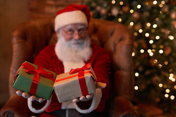 Fototapeta na wymiar Santa Claus sitting in armchair and giving Christmas presents tied with red ribbons to camera