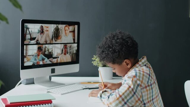 Little Afro-American boy listening to teacher on video call on computer and writing down some notes while taking online class at home during coronavirus lockdown