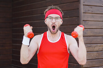 Portrait of funny geek exercising and doing sport training. Crazy athletic man in red sportwear using small dumbbells for bodybuilding.