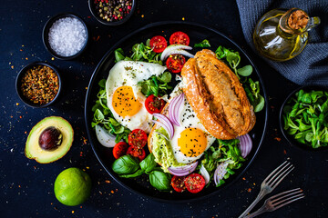 Sunny side up eggs on bun with  fresh green vegetables, mini tomatoes and guacamole on wooden table

