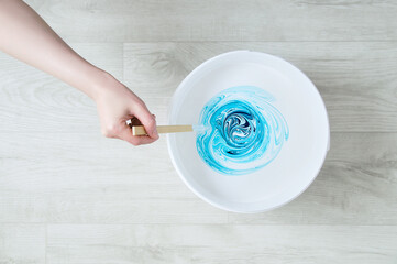 Hand of a Caucasian woman stirring tints of white and blue paint in a bucket on the background of...