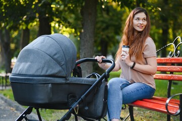 Fototapeta na wymiar Stylish, young mother is sitting on a bench with a baby carriage in a park