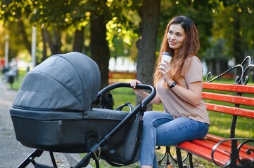 Stylish, young mother is sitting on a bench with a baby carriage in a park
