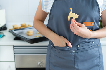 Woman with apron and one hand in pocket holding a halloween cat shaped cookie. Domestic kitchen,....