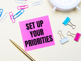 On a light background, a white alarm clock, pink, blue and white paper clips, a golden pen and a pink sticker with the text SET UP YOUR PRIORITIES