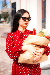 Happy grocery shopper. Portrait of beautiful traditional white woman in red dress holding paper shopping bag full of groceries.