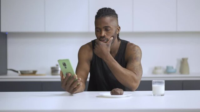 Concept of viewing information at breakfast. Young guy looks thoughtfully at his phone for a glass of milk while sitting at the kitchen table.