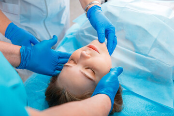 Young woman lies on surgical table before surgery. Surgeon patient portrait on table for rhinoplasty operations with doctors hands in gloves. Surgery or cosmetology beauty treatment inspection