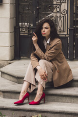 Close up portrait of young beautiful woman with long brunette curly hair posing against building background. sitting on the stairs. bright red high heeled shoes