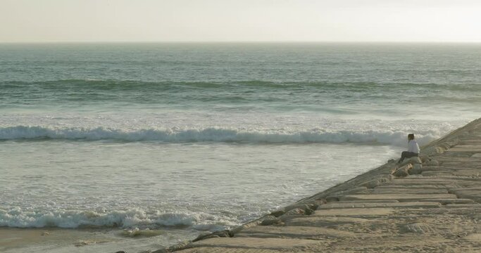 Woman Sitting Near The Edge Of A Sea Wall With Foamy Waves At Figueira da Foz, Coimbra, Portugal. Wide Shot