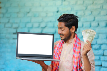 Young indian farmer showing laptop screen and money - obrazy, fototapety, plakaty
