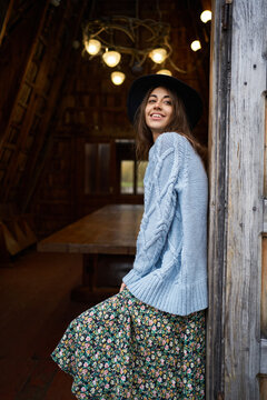 Pretty smiling woman in cozy knitted sweater and hat standing in doorway of wooden house, cozy autumn image