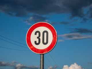 Metal road or traffic sign with number 30 in a red circle, indicating new speed limit against blue sky and clouds