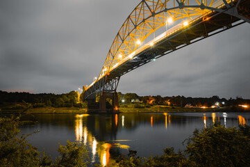 Gold-colored Sagamore Bridge over Cape Cod Canal in Massachusetts at dawn. Glowing bridge lights reflected on the tranquil water. Moody gray clouds passing over the arch.