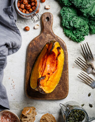 Roasted pumpkin on a wooden cutting board surrounded fall seasonal products. Kale, bread, nuts and seeds. Top view, food frame background.