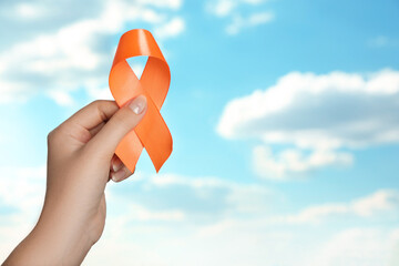 Woman holding orange ribbon against blue sky, closeup with space for text. Multiple sclerosis...