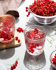 Refreshing drink glass with water, ice and red currants on the table in sunny weather. Peeled currant berries and a bowl of berries on the background