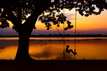 Fototapeta premium A silhouette girl swinging under the silhouette tree with a lake and sunset background