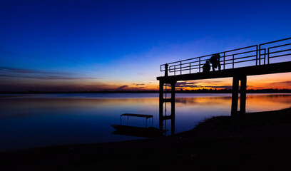 Two silhouette people on the bridge with a sunset background and a boat