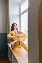 Young business woman meditating lying on the windowsill in the bedroom. Girl listens to calm music. Home comfort zone and wellness concept.