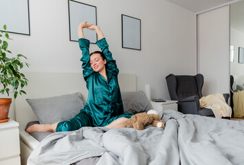 Young woman in silk pajama yawns while sitting in comfortable soft bed.