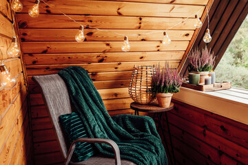 Small natural color wooden cabin balcony with heather flowers, candlelight flame, soft dark green plaid waiting on garden furniture chair. Cute autumn hygge home decor arrangement.