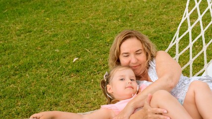 Couple grandmother and granddaughter 3 years old are relaxing and basking in a hammock on a green meadow in their garden on a summer sunny day. Vacation concept, generational relationship, parenting