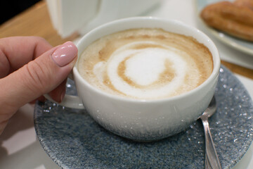 A female hand holds a cup of fresh coffee with milk, a beautiful drawing on top of the foam. Breakfast in a cafe