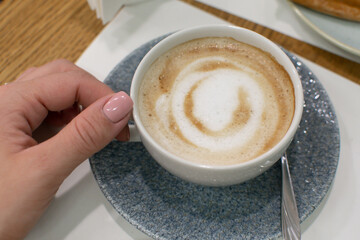 A female hand holds a cup of fresh coffee with milk, a beautiful drawing on top of the foam. Breakfast in a cafe
