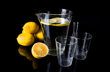 Jug or pitcher of water with lemons and two empty glasses, on black background.