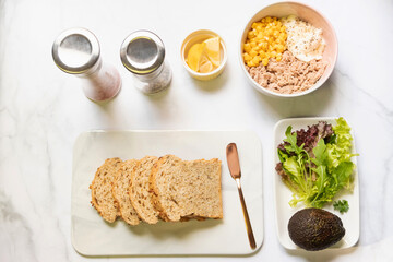 Preparing healthy sandwiches; super grains sliced bread, tuna, corn, avocado, lemon and vegetables on marble cutting board. Top view