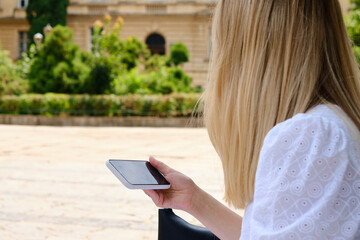 Fototapeta premium A girl on the street is holding a modern smartphone, sending a text message or using an application on her mobile phone. View city map using smartphone. Smartphone close-up