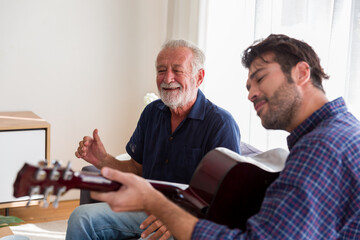 Happy Senior Father sing a song and Adult Son playing guitar on the sofa at home. Elderly father...