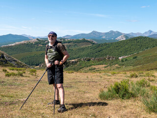 Male hiker with cap, poles and backpack posing on top of a green hill surrounded by a beautiful landscape with the mountains behind