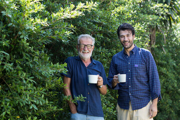 Senior father and adult son walking and talking and drinking coffee together in the garden with...